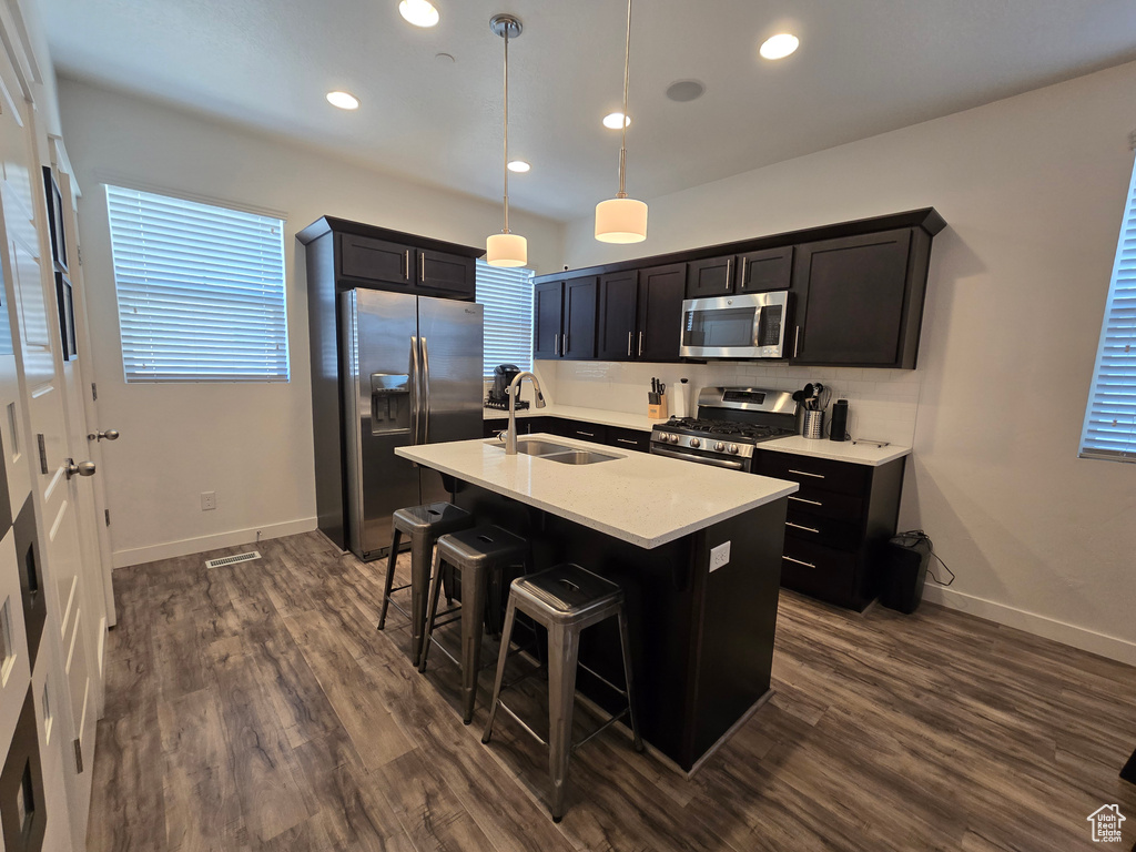 Kitchen featuring an island with sink, stainless steel appliances, decorative light fixtures, dark wood-type flooring, and sink