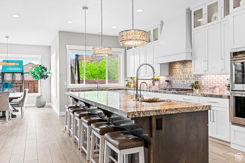 Kitchen featuring light stone counters, backsplash, a center island with sink, custom exhaust hood, and decorative light fixtures