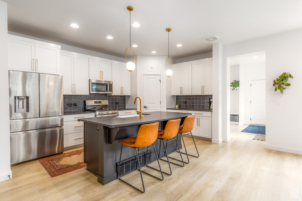 Kitchen with hanging light fixtures, appliances with stainless steel finishes, a center island with sink, light hardwood / wood-style floors, and tasteful backsplash