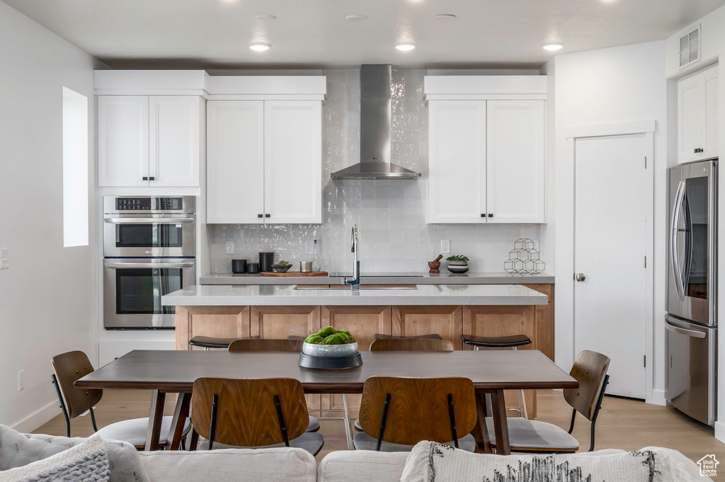 Kitchen featuring white cabinetry, appliances with stainless steel finishes, light hardwood / wood-style floors, tasteful backsplash, and wall chimney exhaust hood