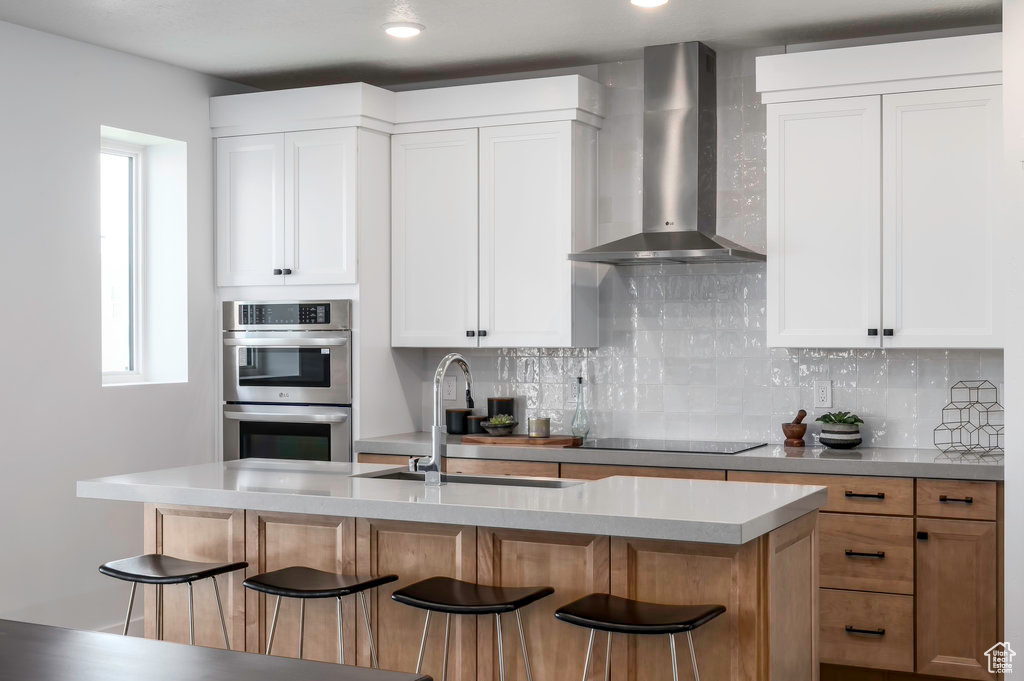 Kitchen featuring white cabinets, backsplash, a breakfast bar area, double oven, and wall chimney range hood