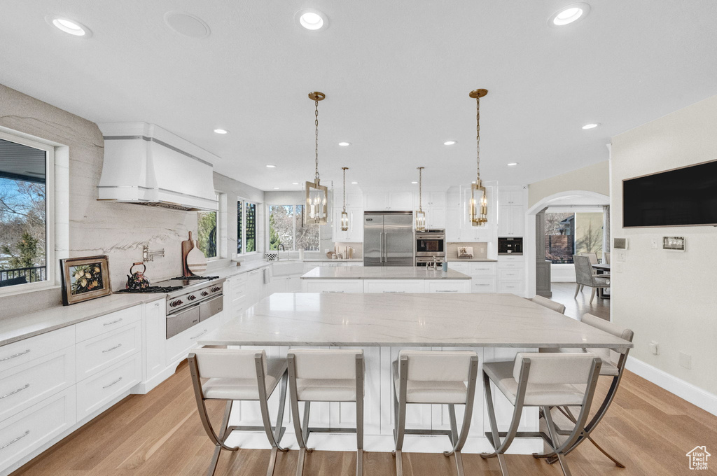 Kitchen featuring appliances with stainless steel finishes, light hardwood / wood-style floors, premium range hood, white cabinets, and pendant lighting