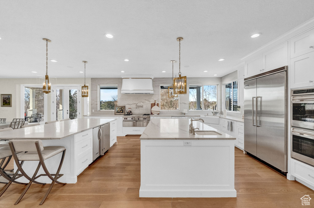 Kitchen with custom exhaust hood, a kitchen island with sink, white cabinetry, and stainless steel appliances