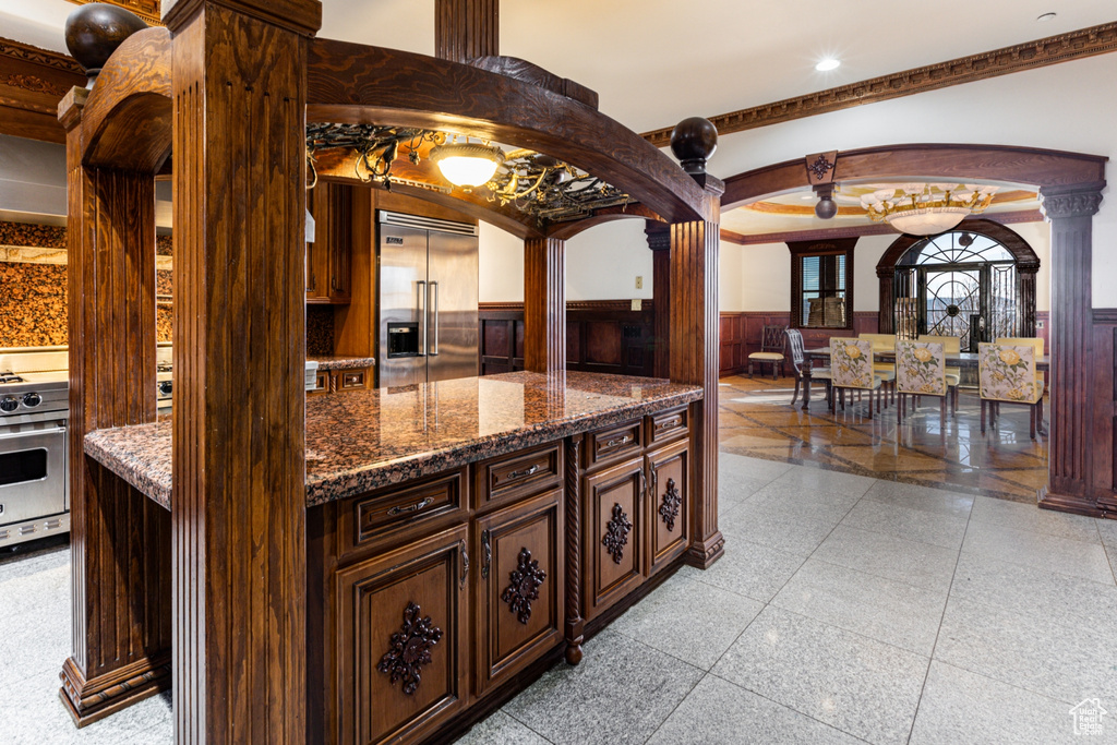 Kitchen featuring light tile floors, dark stone counters, crown molding, stainless steel appliances, and ornate columns