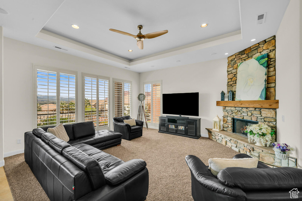 Living room featuring light carpet, a tray ceiling, a stone fireplace, and ceiling fan