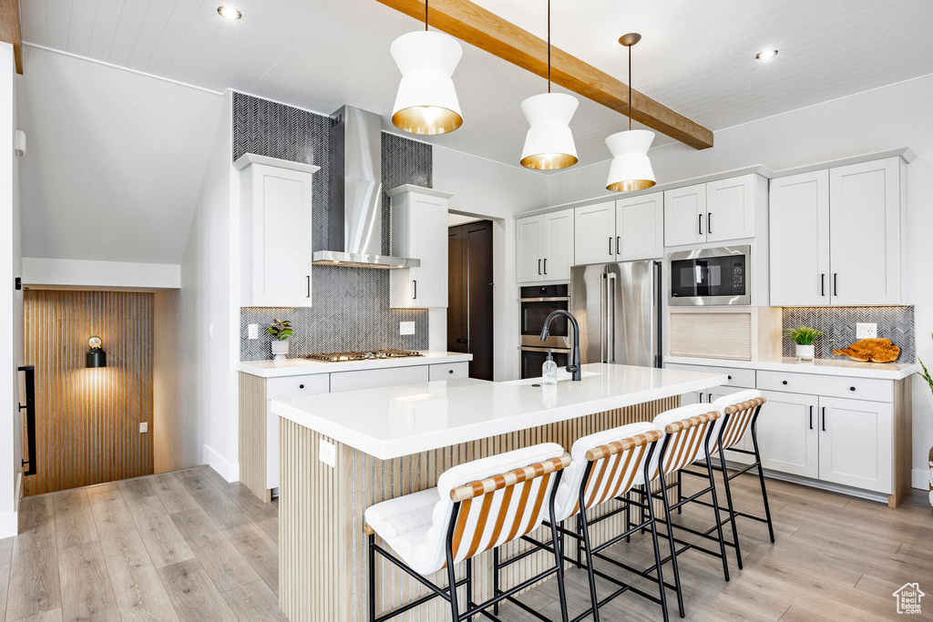 Kitchen featuring an island with sink, appliances with stainless steel finishes, backsplash, light hardwood / wood-style floors, and wall chimney range hood