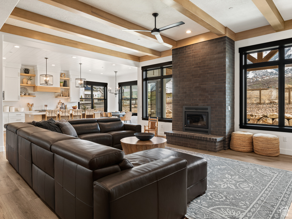 Living room featuring a brick fireplace, beamed ceiling, ceiling fan with notable chandelier, and light hardwood / wood-style flooring