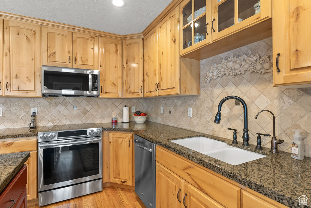Kitchen with appliances with stainless steel finishes, tasteful backsplash, dark stone countertops, light hardwood / wood-style floors, and sink
