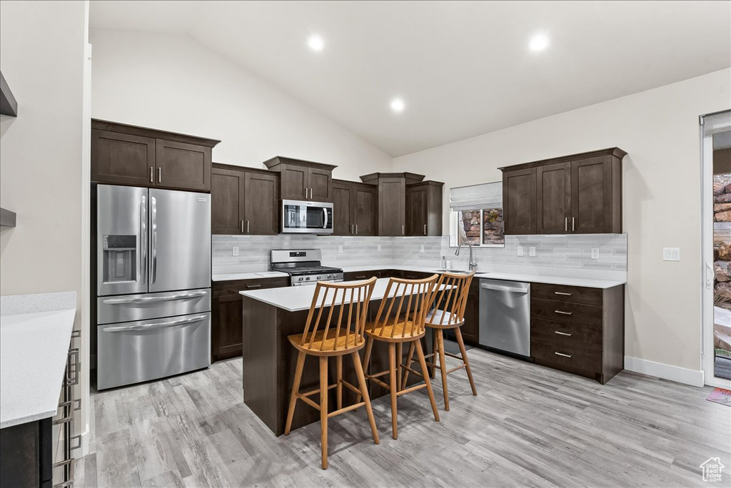 Kitchen with appliances with stainless steel finishes, high vaulted ceiling, light wood-type flooring, tasteful backsplash, and a breakfast bar area