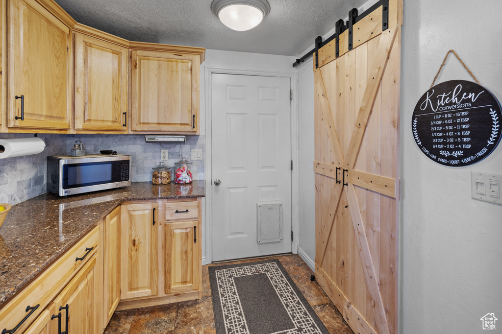 Kitchen with dark stone counters, tasteful backsplash, a barn door, light brown cabinetry, and dark tile flooring