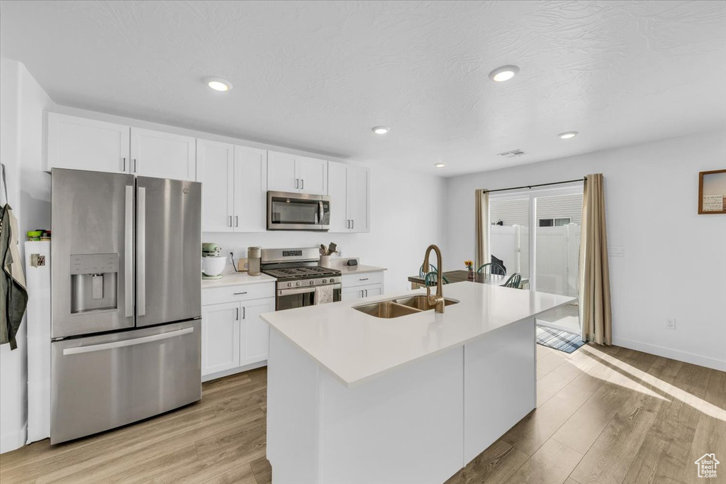 Kitchen with an island with sink, white cabinetry, appliances with stainless steel finishes, light wood-type flooring, and sink