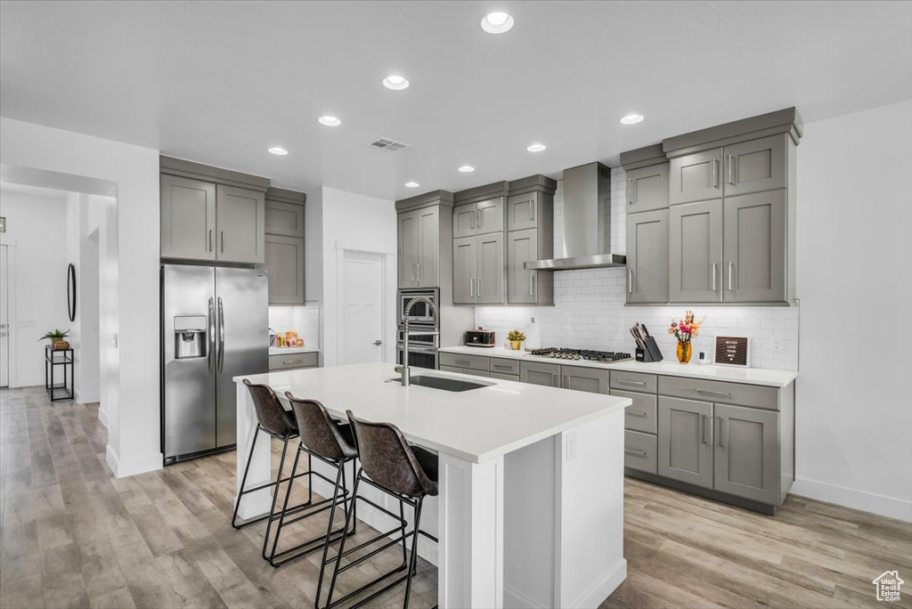 Kitchen featuring appliances with stainless steel finishes, an island with sink, light hardwood / wood-style flooring, wall chimney range hood, and tasteful backsplash