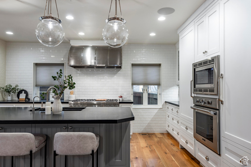 Kitchen featuring hanging light fixtures, light hardwood / wood-style flooring, white cabinetry, wall chimney range hood, and stainless steel appliances