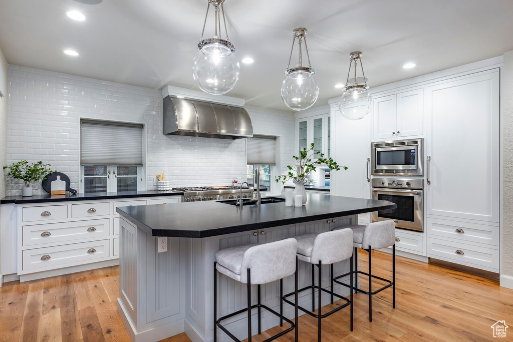 Kitchen featuring wall chimney exhaust hood, light hardwood / wood-style flooring, backsplash, appliances with stainless steel finishes, and an island with sink