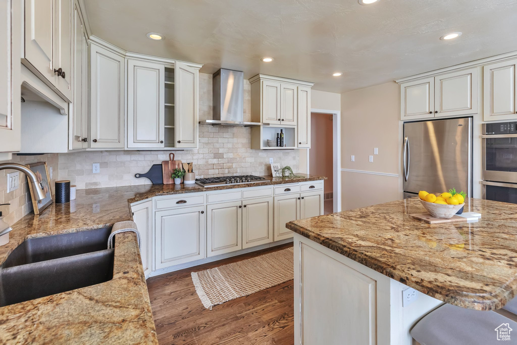 Kitchen with backsplash, stainless steel appliances, light stone counters, wall chimney range hood, and dark hardwood / wood-style flooring