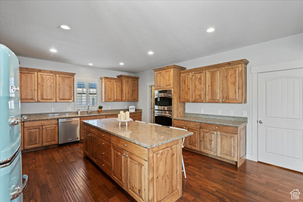 Kitchen with stainless steel appliances, a center island, light stone countertops, dark wood-type flooring, and sink