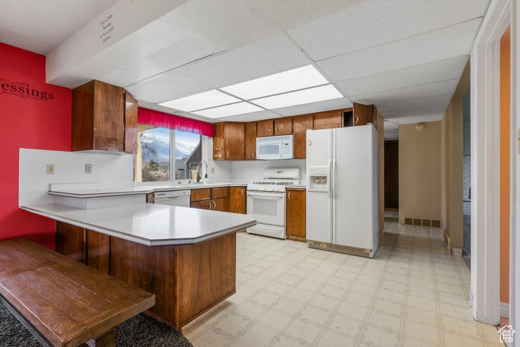 Kitchen with kitchen peninsula, white appliances, sink, light tile floors, and a paneled ceiling
