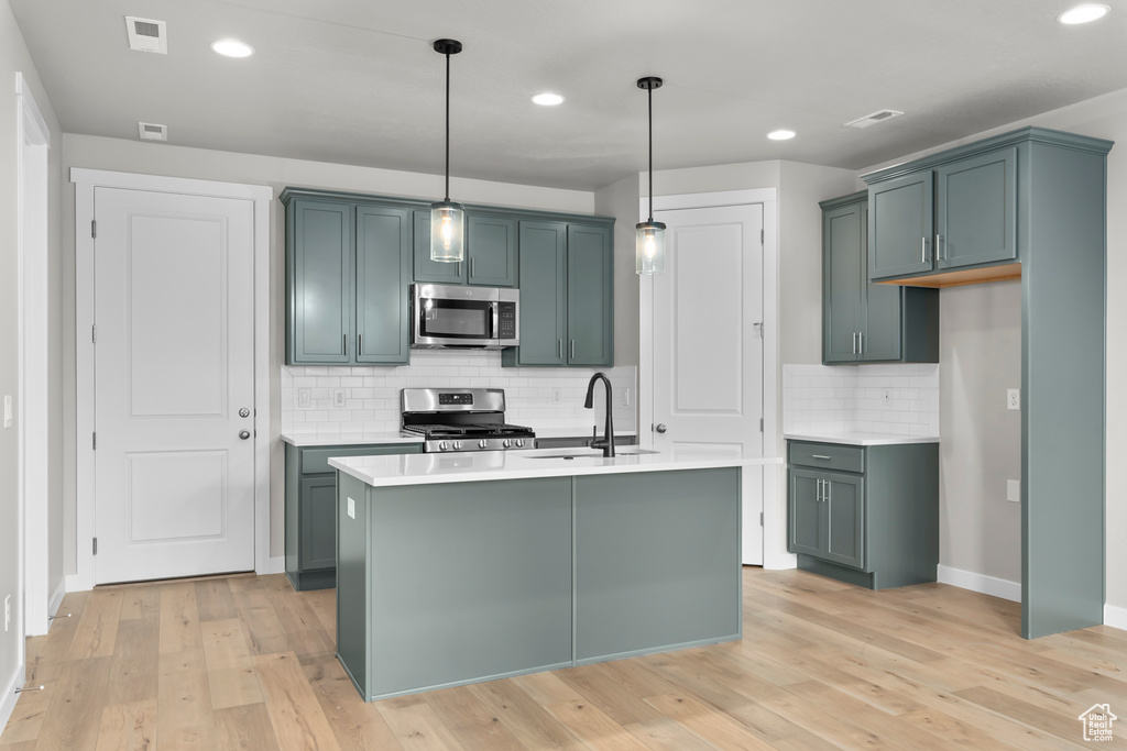 Kitchen with pendant lighting, appliances with stainless steel finishes, light hardwood / wood-style flooring, a center island with sink, and tasteful backsplash