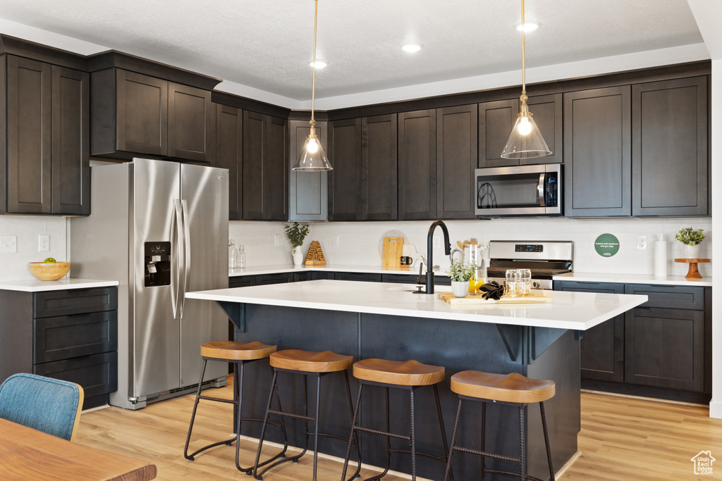 Kitchen with decorative light fixtures, a center island with sink, appliances with stainless steel finishes, dark brown cabinetry, and light wood-type flooring