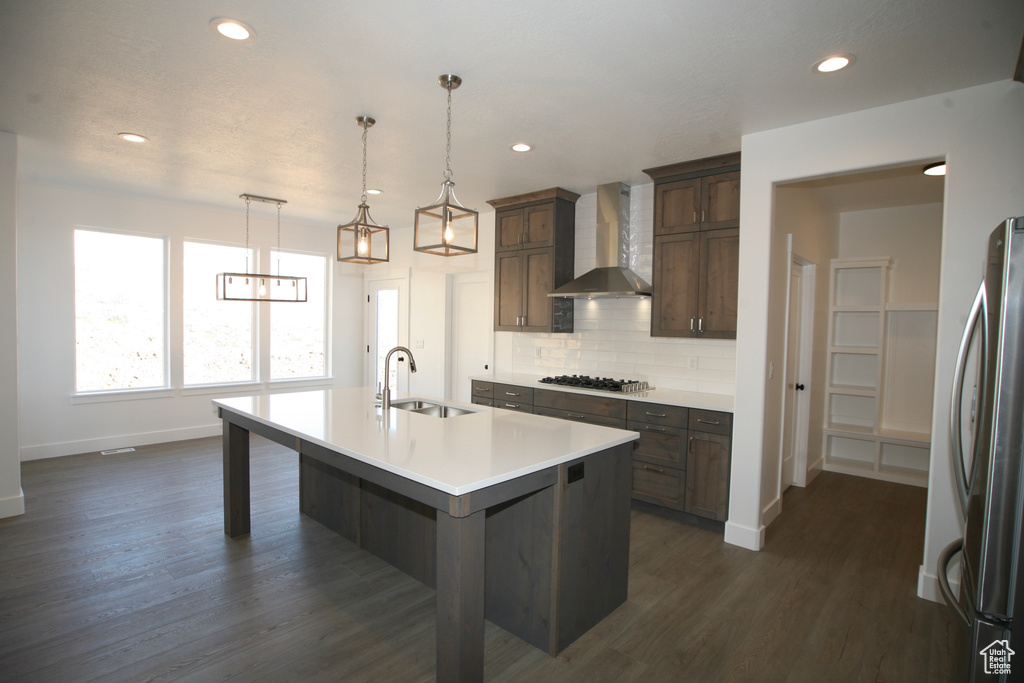 Kitchen with decorative light fixtures, dark wood-type flooring, sink, an island with sink, and wall chimney range hood