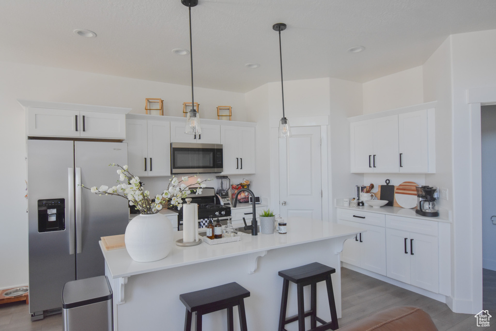Kitchen featuring an island with sink, light hardwood / wood-style floors, stainless steel appliances, decorative light fixtures, and white cabinetry