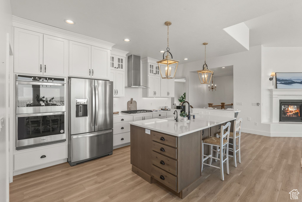 Kitchen with light hardwood / wood-style floors, a center island with sink, wall chimney exhaust hood, white cabinetry, and appliances with stainless steel finishes