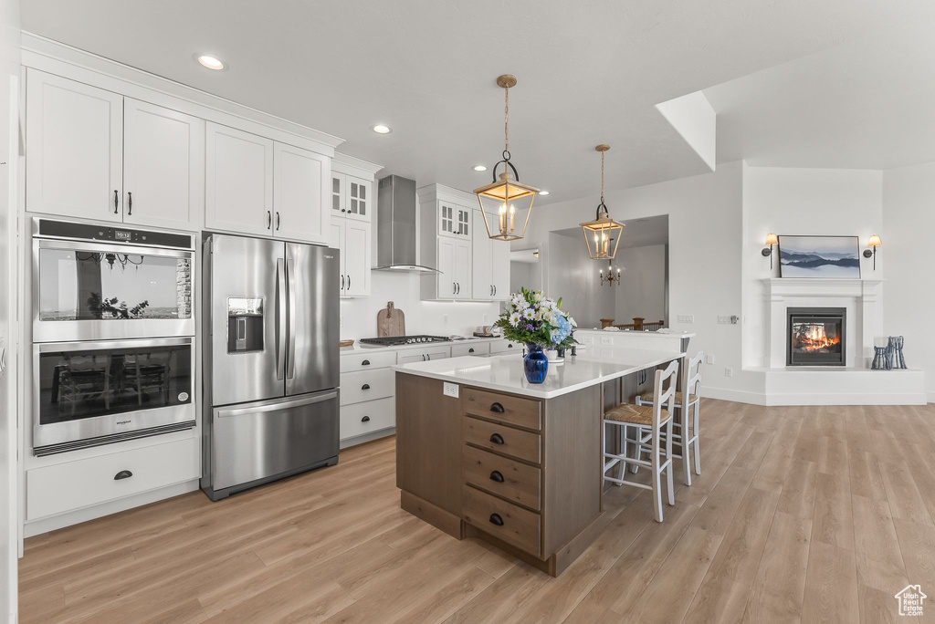 Kitchen with appliances with stainless steel finishes, white cabinets, light hardwood / wood-style flooring, a center island, and wall chimney range hood
