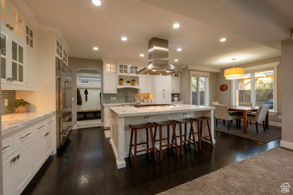 Kitchen with decorative light fixtures, dark wood-type flooring, white cabinetry, island exhaust hood, and backsplash