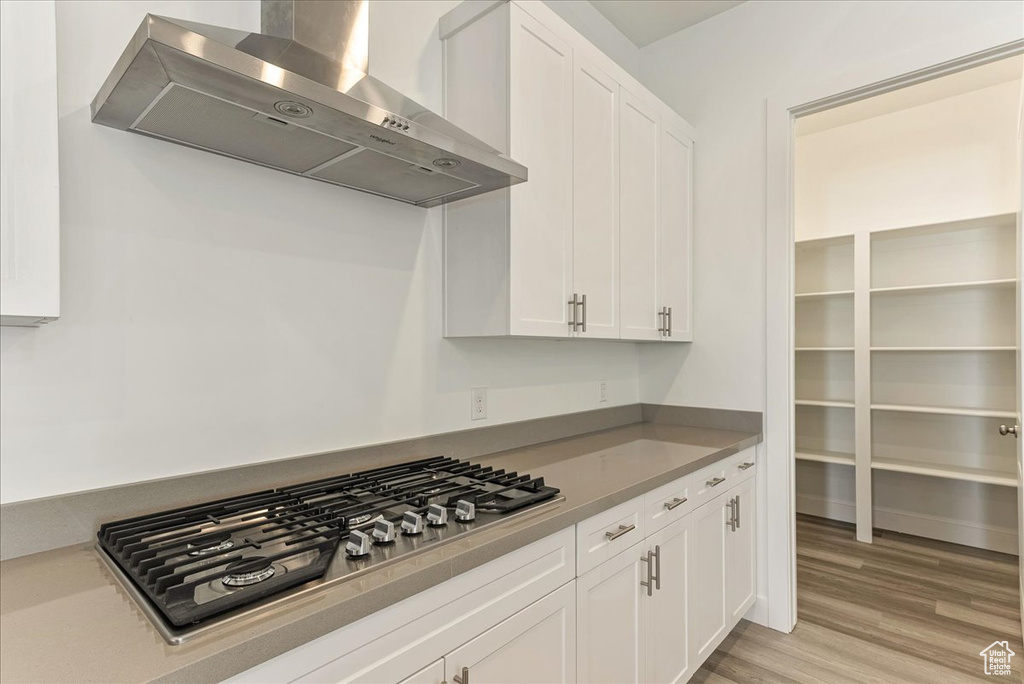 Kitchen with wall chimney range hood, stainless steel gas cooktop, white cabinetry, and light wood-type flooring