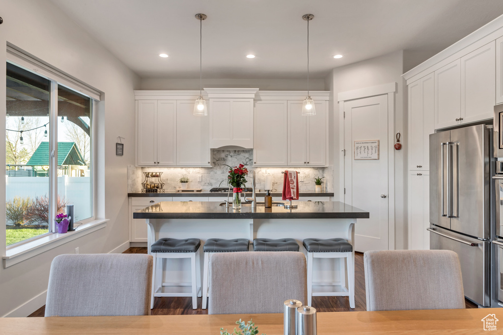 Kitchen with high quality fridge, hanging light fixtures, white cabinets, and a kitchen island with sink