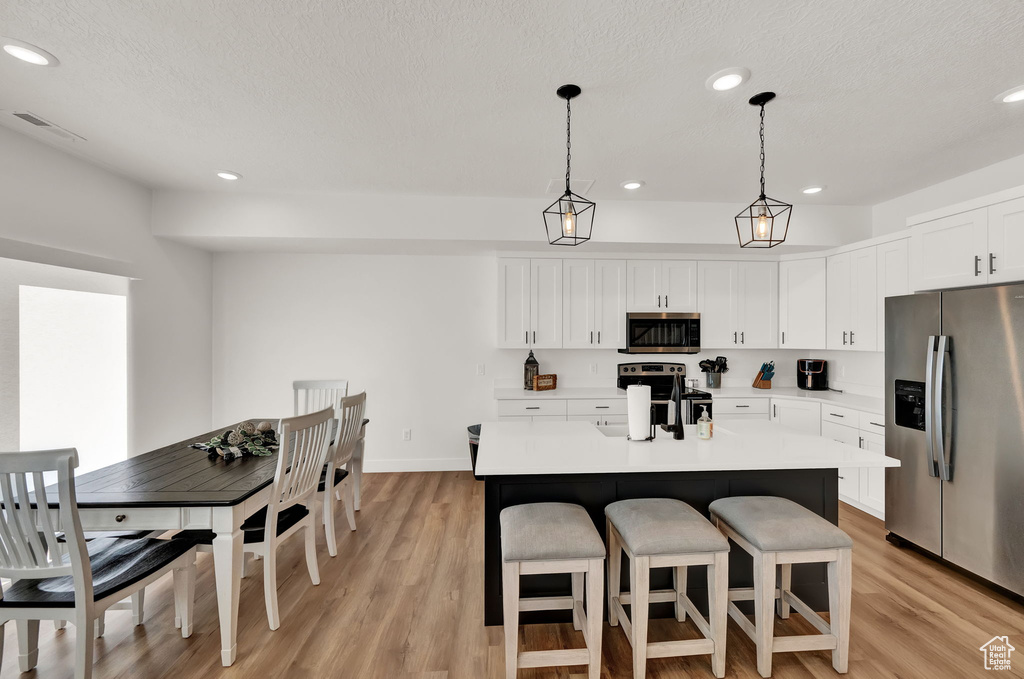 Kitchen featuring appliances with stainless steel finishes, an island with sink, light wood-type flooring, white cabinets, and pendant lighting