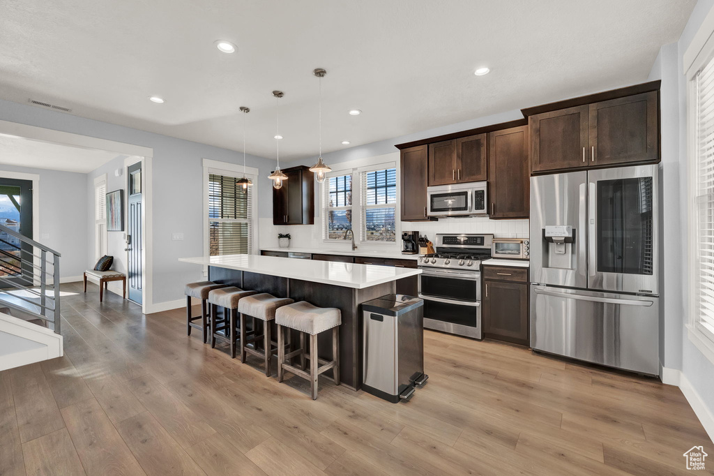 Kitchen with appliances with stainless steel finishes, a kitchen island, decorative light fixtures, dark brown cabinetry, and light hardwood / wood-style flooring