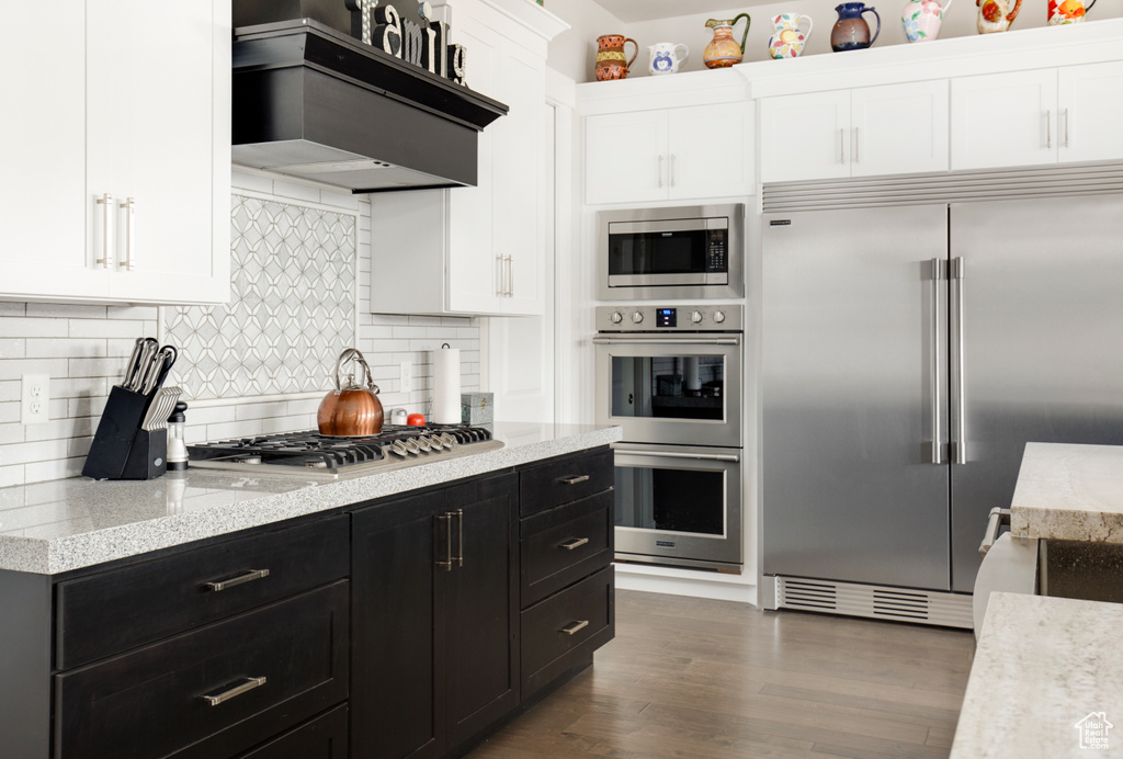 Kitchen featuring built in appliances, custom exhaust hood, dark hardwood / wood-style flooring, backsplash, and light stone counters
