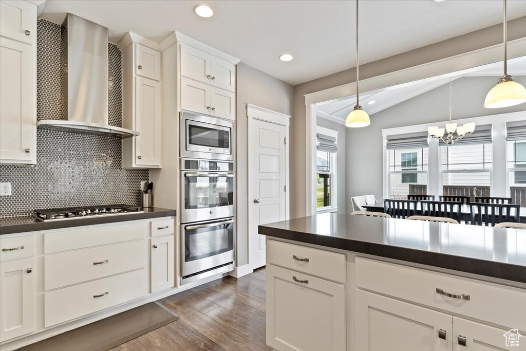 Kitchen featuring dark wood-type flooring, a notable chandelier, stainless steel appliances, decorative light fixtures, and wall chimney exhaust hood