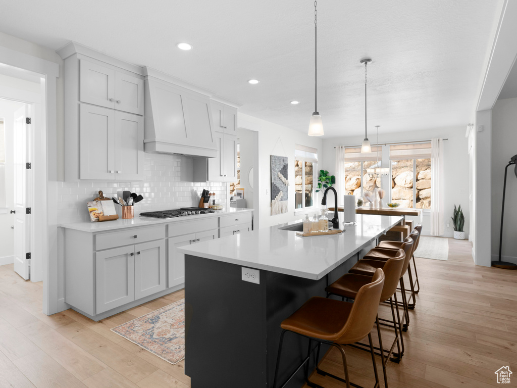 Kitchen featuring a kitchen island with sink, custom exhaust hood, sink, light wood-type flooring, and tasteful backsplash