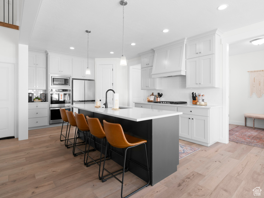 Kitchen featuring premium range hood, a center island with sink, appliances with stainless steel finishes, light wood-type flooring, and sink