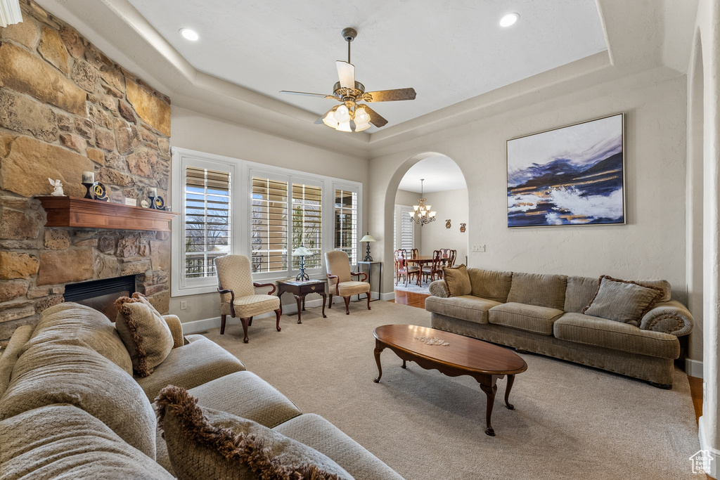 Living room with a stone fireplace, light carpet, a tray ceiling, and ceiling fan with notable chandelier