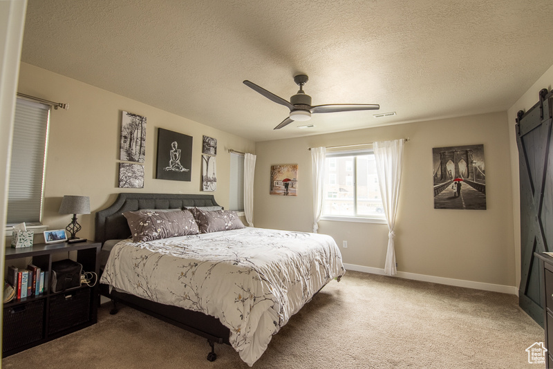 Bedroom with a barn door, ceiling fan, light carpet, and a textured ceiling