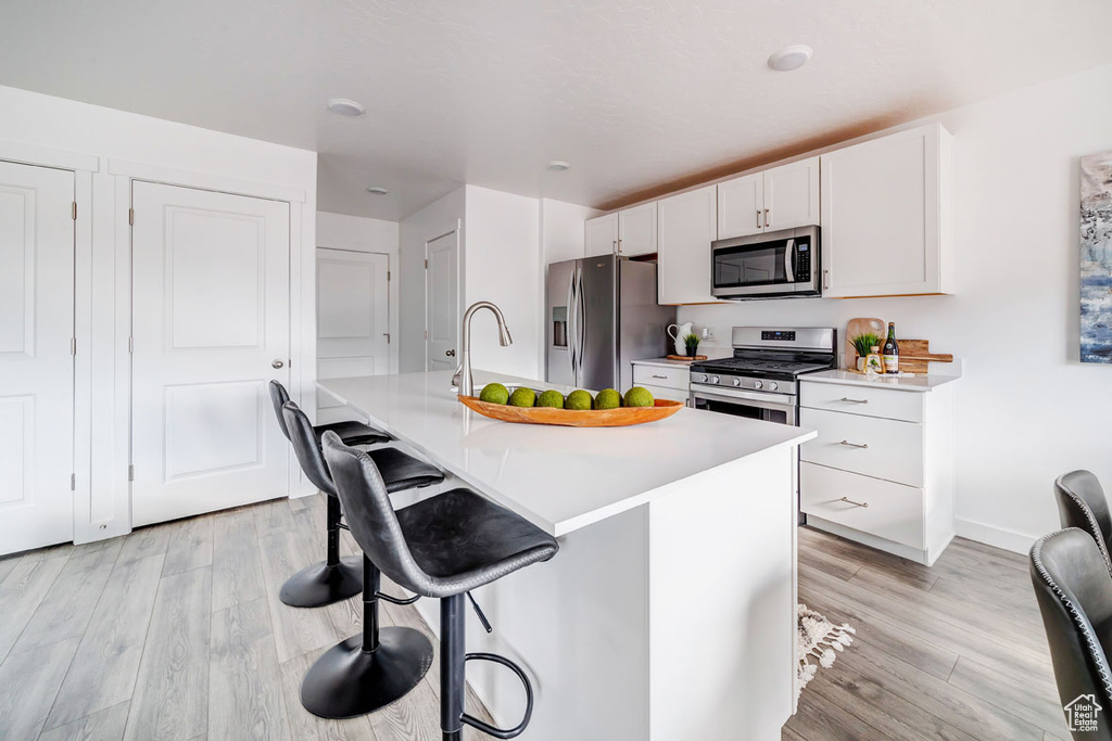 Kitchen featuring white cabinetry, a kitchen bar, an island with sink, stainless steel appliances, and light hardwood / wood-style flooring
