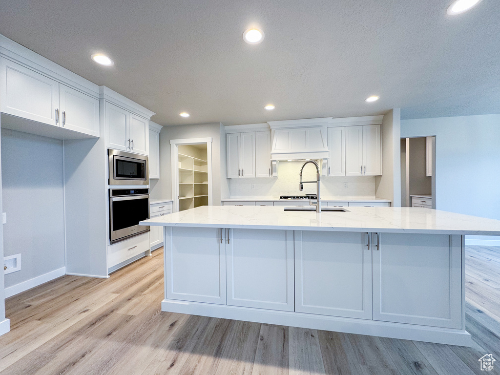 Kitchen featuring an island with sink, premium range hood, light hardwood / wood-style floors, stainless steel appliances, and white cabinetry