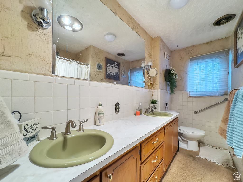 Bathroom with toilet, dual bowl vanity, backsplash, a textured ceiling, and tile walls