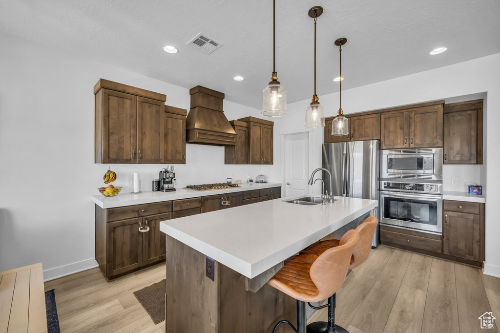 Kitchen featuring appliances with stainless steel finishes, pendant lighting, custom exhaust hood, a kitchen island with sink, and sink