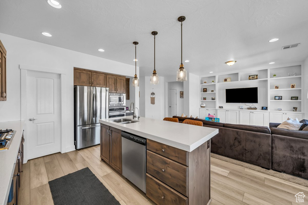 Kitchen featuring light hardwood / wood-style flooring, appliances with stainless steel finishes, hanging light fixtures, a kitchen island with sink, and sink