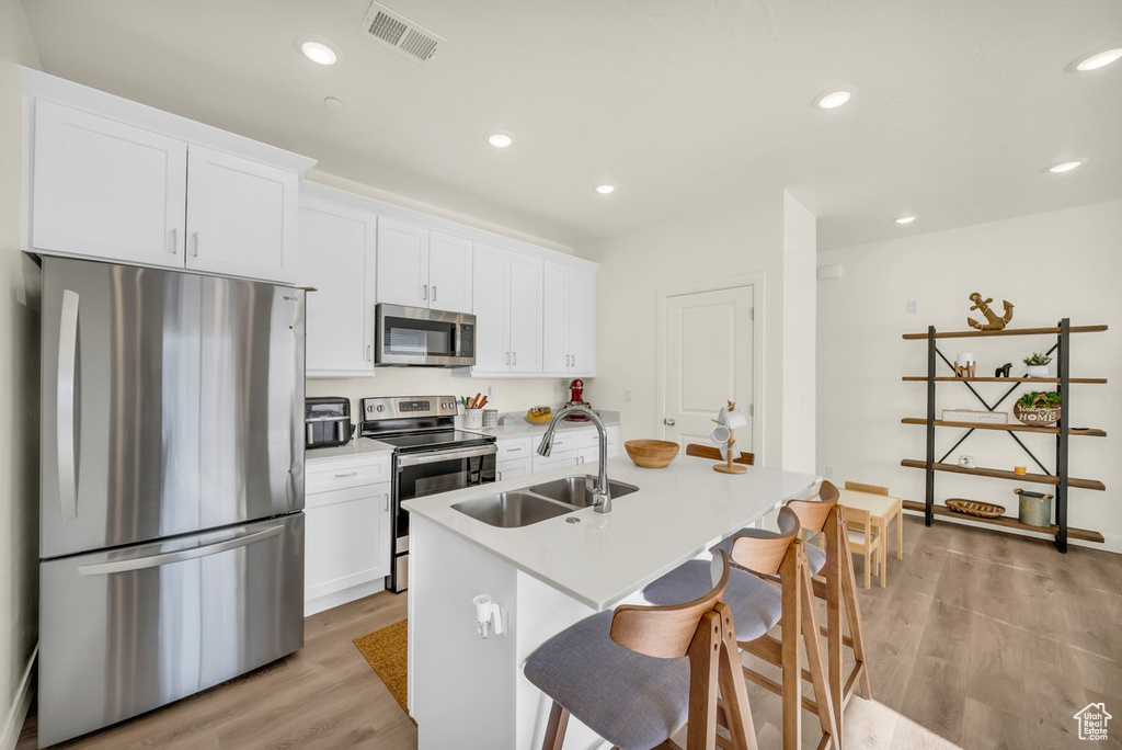 Kitchen with stainless steel appliances, sink, white cabinetry, light hardwood / wood-style flooring, and a kitchen island with sink
