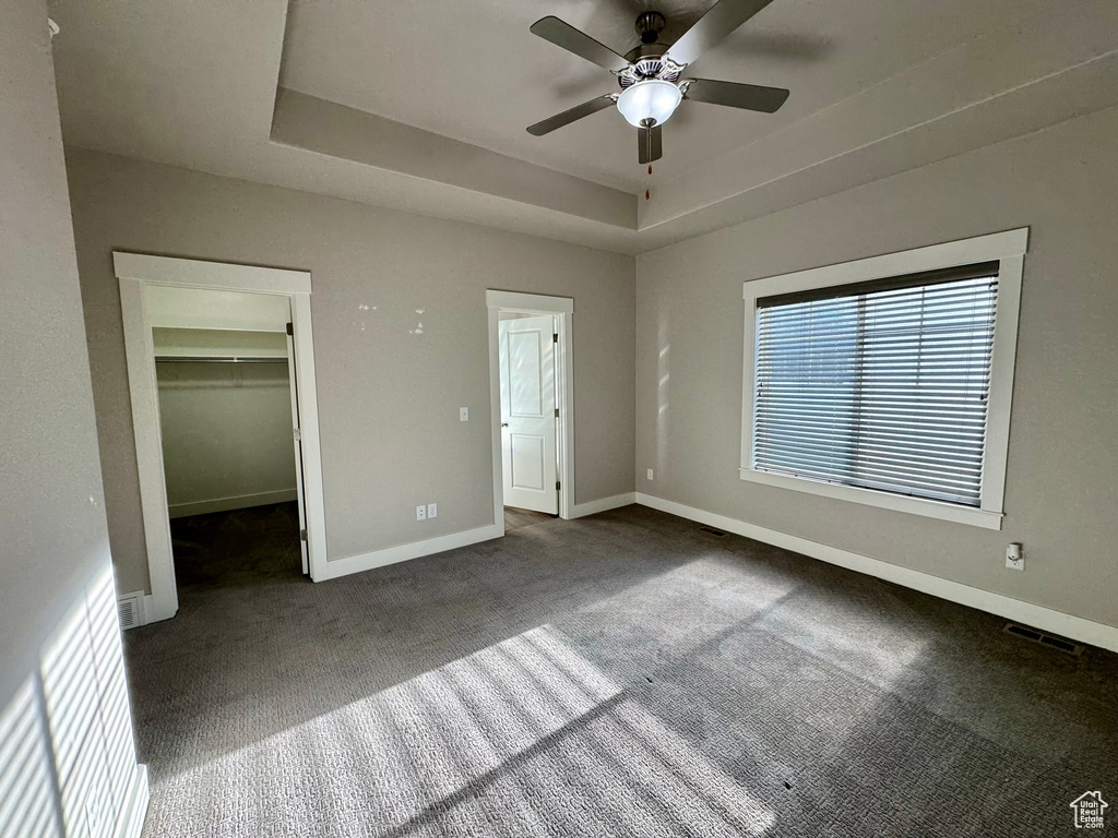 Unfurnished bedroom with dark carpet, a closet, a spacious closet, a raised ceiling, and ceiling fan
