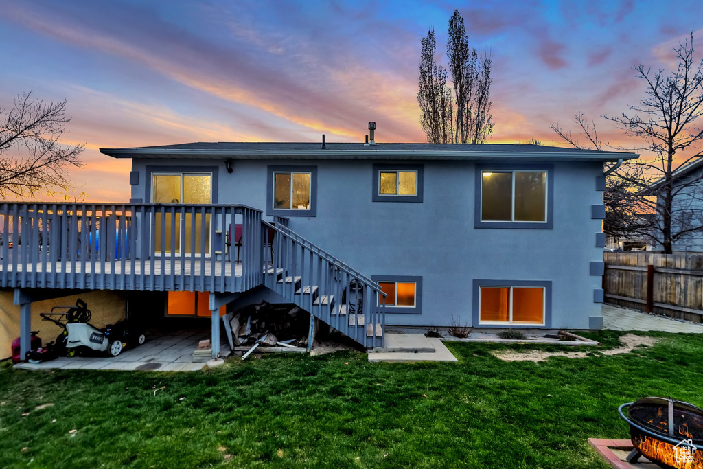 Back house at dusk featuring a wooden deck, a patio area, a lawn, and an outdoor fire pit