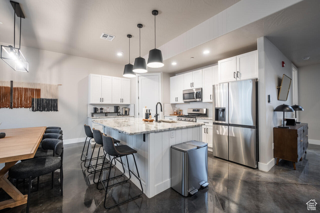 Kitchen featuring hanging light fixtures, appliances with stainless steel finishes, an island with sink, white cabinets, and light stone counters