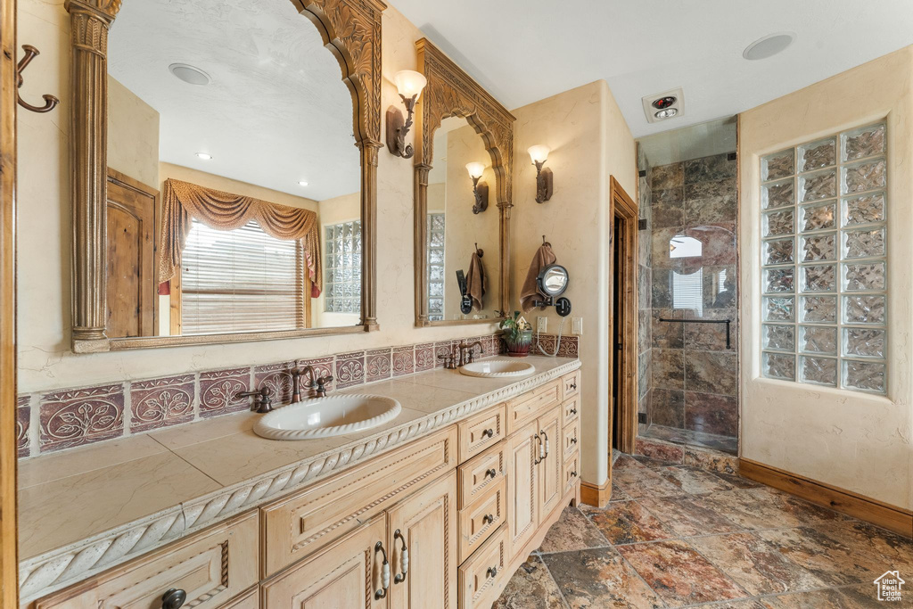 Bathroom featuring a shower with door, double sink, vanity with extensive cabinet space, backsplash, and tile floors