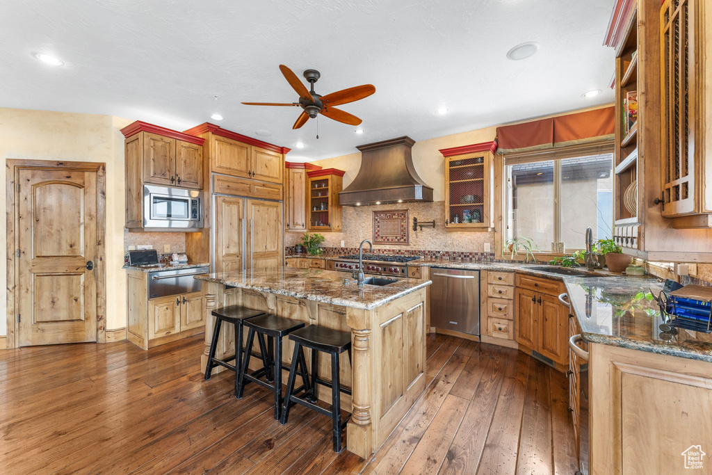 Kitchen featuring appliances with stainless steel finishes, a center island with sink, a breakfast bar area, custom range hood, and ceiling fan