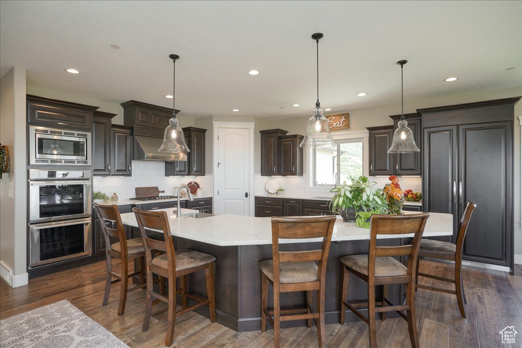 Kitchen featuring dark hardwood / wood-style floors, a kitchen island with sink, pendant lighting, stainless steel microwave, and backsplash
